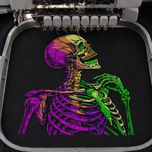 Könnte beinhalten: Ein gesticktes Design eines farbenfrohen Skeletts auf schwarzem Stoff. Der Schädel ist gelb und orange, der Brustkorb und die Arme sind lila und grün, und der Hintergrund ist schwarz. Das Design wird von einer Stickmaschine erstellt.