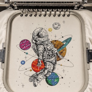 May include: Embroidered design of an astronaut floating in space, surrounded by colorful planets and stars. The astronaut is detailed in black and white, with planets in shades of red, orange, blue, and purple. The design is set within an embroidery hoop.