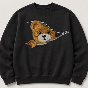 May include: Black crewneck sweatshirt featuring an embroidered teddy bear design. The bear is peeking out from a zipper, with brown fur, a white face, and a black nose and eyes. The zipper is white.