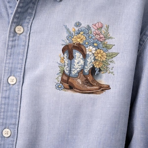 May include: Light blue denim shirt featuring an embroidered design of brown cowboy boots with blue and white detailing, surrounded by colorful flowers. The shirt has a button-down collar and buttoned cuffs.