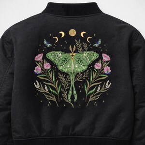 May include: Black bomber jacket featuring an embroidered design on the back. The design includes a large green moth, crescent moons, butterflies, and floral elements in shades of pink, green, and gold.