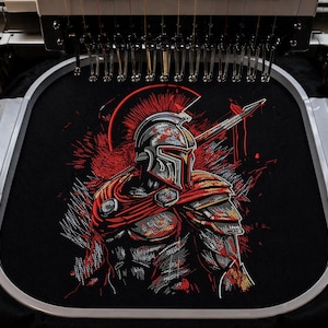 May include: Black t-shirt featuring an embroidered Spartan warrior design. The warrior is depicted in a helmet and armour, holding a sword, with red and grey accents. The shirt is in an embroidery machine, ready for the next step.