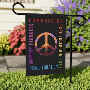 May include: A black garden flag with a peace sign in the center. The peace sign is filled with a colorful sunset and flowers. The words "COMPASSION", "HOPE", "INCLUSION", "LOVE", "DIVERSITY", "KINDNESS", "PEACE", "EQUALITY" are written around the peace sign in a rainbow of colors.