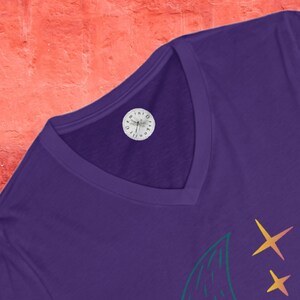 May include: Purple V-neck t-shirt with a green and yellow crescent moon and stars graphic. The shirt has a small white label with a dragonfly and the text "Dreamin' Butterfly Co."