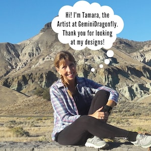 May include: A woman wearing a plaid shirt and black pants sits on a rock wall in front of a colorful mountain landscape. She is smiling and looking at the camera. A thought bubble above her head reads "Hi! I'm Tamara, the Artist at GeminiDragonfly. Thank you for looking at my designs!"