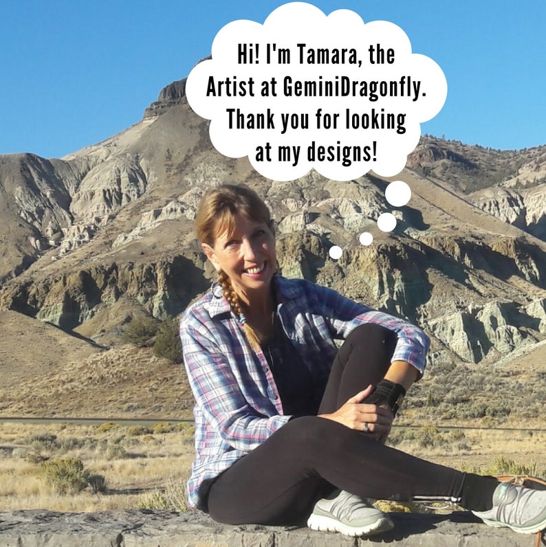 May include: A woman wearing a plaid shirt and black pants sits on a rock wall in front of a colorful mountain landscape. She is smiling and looking at the camera. A thought bubble above her head reads "Hi! I'm Tamara, the Artist at GeminiDragonfly. Thank you for looking at my designs!"