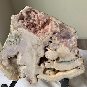 May include: A large, irregularly shaped geode with a rough, textured exterior. The interior reveals a cluster of pink and white crystals, with hints of green and brown. The geode is displayed on a black stand, showcasing its natural beauty.