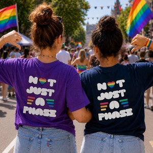 May include: A purple t-shirt and a navy blue t-shirt, both featuring the white text "NOT JUST A PHASE" with a rainbow heart icon and colorful horizontal bars. The shirts are worn with light blue denim shorts, showcasing a casual, everyday look.
