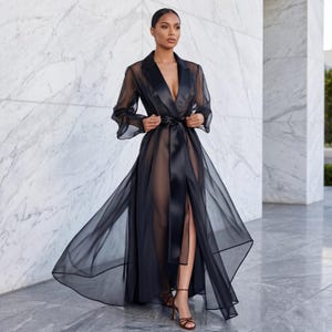 May include: A sheer black robe with a satin lapel and belt. The long, flowing robe has a high slit and is worn over a matching outfit. The model is wearing brown high heels. The background is a marble wall.