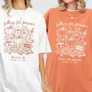 May include: Two oversized t-shirts, one white with a terracotta design and one terracotta with a white design. Both feature the text 'falling for forever', 'Miami FL 2026', and illustrations of pumpkins, leaves, a mug, a drink, cake, and a scarf.