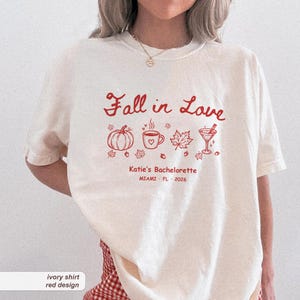 May include: An ivory t-shirt featuring a red graphic design with the text Fall in Love, Katie's Bachelorette, and MIAMI FL 2026. The print includes illustrations of a pumpkin, a coffee mug, a maple leaf, and a martini glass with small leaves.