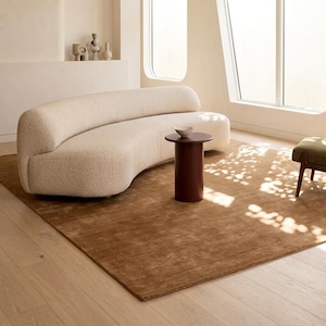 May include: A curved cream-coloured sofa with a textured bouclé fabric, paired with a cylindrical dark brown side table and a small bowl. The furniture sits on a large, solid brown area rug in a room with light-coloured walls and large windows.