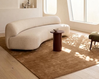 May include: A curved cream-coloured sofa with a textured bouclé fabric, paired with a cylindrical dark brown side table and a small bowl. The furniture sits on a large, solid brown area rug in a room with light-coloured walls and large windows.