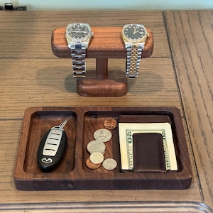 May include: A wooden valet tray with a watch stand. Two watches with silver and gold bands rest on the stand. The tray holds a car key, coins, a wallet, and cash. The wood has a rich, dark finish.