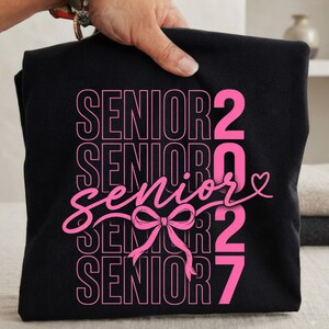 May include: A black t-shirt featuring the word SENIOR repeated in a stacked pink outline font. The word senior is written in a pink cursive script across the center, accented by a pink bow and heart. The number 2027 is printed in pink.