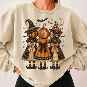 May include: A beige crewneck sweatshirt featuring a graphic of three pumpkin-headed figures wearing witch hats and dresses with bows. The design includes small stars, bats, and a crescent moon, rendered in shades of orange, black, and cream.