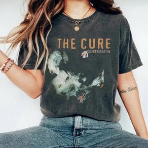 May include: A charcoal gray short-sleeve t-shirt featuring the text THE CURE and DISINTEGRATION in a tan font. The graphic includes a faded, artistic portrait of a person surrounded by abstract floral elements in muted green and white tones.