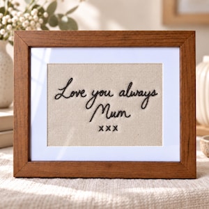 Può includere: Una cornice rettangolare in legno contenente un tessuto beige con il testo ricamato in nero Love you always Mum seguito da tre simboli x. La cornice presenta un bordo bianco che circonda il pannello centrale in tessuto.