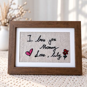 Könnte beinhalten: Ein rechteckiger Holzrahmen mit einem hellbeigen Stoffstück, auf dem der schwarze Sticktext I love you Mommy Love, lily zu sehen ist. Die Stickerei enthält zudem ein kleines rosa Herz und eine rote Blume mit grünem Stiel auf dem Stoff.