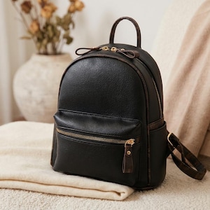 Puede incluir: Mochila negra de cuero sintético con forma redondeada, con un bolsillo frontal con cremallera y herrajes dorados. La mochila tiene ribetes marrones y un asa superior. Está sobre una manta color crema.