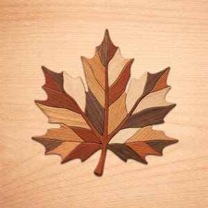 May include: A decorative maple leaf shaped object constructed from multiple segments of wood in varying shades of brown, tan, and cream. The piece features a central stem and distinct pointed lobes, showcasing the natural grain patterns of the wood.