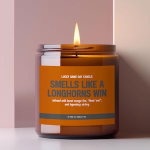 Puede incluir: Una vela encendida en un frasco de vidrio marrón con una etiqueta naranja. La etiqueta dice: LUCKY GAME DAY CANDLE, SMELLS LIKE A LONGHORNS WIN, infused with burnt orange fire, "Hook 'em!", and legendary victory, BY CARA CO. CANDLE | 255g.