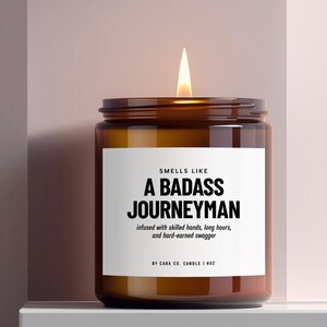 May include: A lit candle in an amber glass jar with a white label that reads: SMELLS LIKE A BADASS JOURNEYMAN, infused with skilled hands, long hours, and hard-earned swagger, BY CARA CO. CANDLE | 9OZ. The candle features a single burning wick.