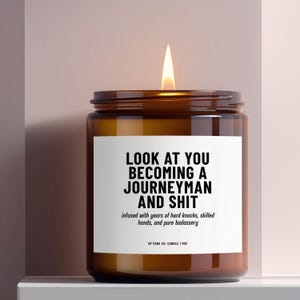 May include: A lit candle in a brown glass jar with a white label. The label reads "LOOK AT YOU BECOMING A JOURNEYMAN AND SHIT" with additional text. The candle is on a white shelf against a pink background.