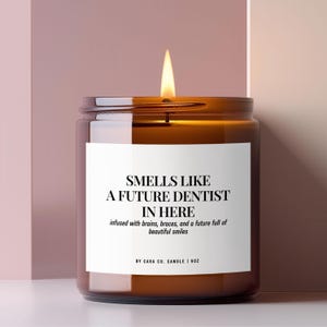 May include: A lit candle in a brown glass jar with a white label that reads: SMELLS LIKE A FUTURE DENTIST IN HERE, infused with brains, braces, and a future full of beautiful smiles, BY CARA CO. CANDLE | 9OZ. The candle has a single burning wick.