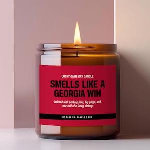 Puede incluir: Una vela encendida en un frasco de vidrio marrón con una etiqueta roja. La etiqueta dice: LUCKY GAME DAY CANDLE, SMELLS LIKE A GEORGIA WIN, infused with barking fans, big plays, and one hell of a Dawg victory, BY CARA CO. CANDLE | 9OZ.