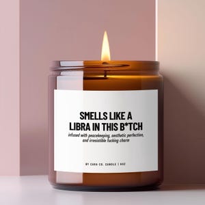 May include: A brown glass candle jar with a lit wick and a white label featuring black text that reads: SMELLS LIKE A LIBRA IN THIS B*TCH, infused with peacekeeping, aesthetic perfection, and irresistible fucking charm, BY CARA CO. CANDLE | 9OZ.