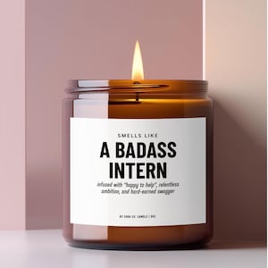 May include: A lit candle in an amber glass jar with a white label that reads: SMELLS LIKE A BADASS INTERN, infused with "happy to help", relentless ambition, and hard-earned swagger, BY CARA CO. CANDLE | 9OZ. The candle has a single burning wick.