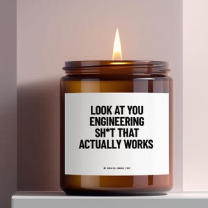May include: A lit candle in a brown glass jar with a black lid. The white label reads "LOOK AT YOU ENGINEERING SH*T THAT ACTUALLY WORKS". The candle is on a white shelf against a neutral background.