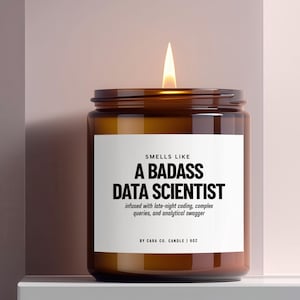 May include: A lit candle in a brown glass jar with a white label that reads: SMELLS LIKE A BADASS DATA SCIENTIST, infused with late-night coding, complex queries, and analytical swagger, BY CARA CO. CANDLE | 9OZ. The candle has a single burning wick.