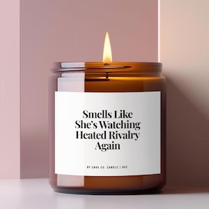 May include: A brown glass jar candle with a lit wick and a white label featuring the black text: Smells Like She's Watching Heated Rivalry Again, BY CARA CO. CANDLE | 9OZ. The candle is cylindrical with a smooth finish and a visible flame at the top.