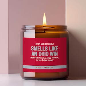 May include: A lit candle in a brown glass jar with a red label. The label text reads: LUCKY GAME DAY CANDLE, SMELS LIKE AN OHIO WIN, infused with Horseshoe energy, rival tears, and pure buckeye swagger, BY CARA CO. CANDLE | 9OZ.