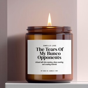 Puede incluir: Una vela encendida en un frasco de vidrio ámbar con una etiqueta blanca que tiene el texto negro: SMELLS LIKE The Tears Of My Bunco Opponents, infused with dice mastery, sharp counting, and crushing victories, BY CARA CO. CANDLE | 255g.