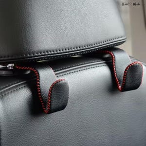 Peut inclure: Un crochet de repose-tête de siège de voiture en similicuir noir avec une surface texturée et des coutures rouges contrastantes sur les bords. Le crochet est conçu pour se fixer aux tiges métalliques de l'appui-tête d'un véhicule.