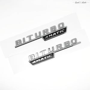May include: Two silver "BITURBO 4MATIC" emblems. The emblems are on a white background. The text is in a bold, sans-serif font. The "4MATIC" text is in a black rectangle. The top right corner has the words "Hand Made" in a script font.