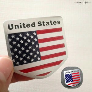 Puede incluir: Un emblema plateado en forma de escudo con el diseño de la bandera de los Estados Unidos. La bandera tiene rayas rojas y blancas con un campo azul de estrellas blancas. Las palabras "United States" están impresas en negro en la parte superior. Se ve una versión más pequeña.