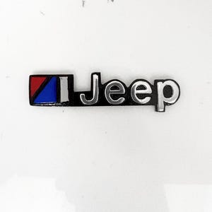 Puede incluir: Un emblema de Jeep con letras plateadas sobre un fondo negro. A la izquierda del texto hay un icono rectangular dividido en tres secciones: un triángulo rojo, un triángulo azul y una barra vertical plateada, todo con un borde negro.