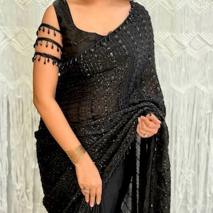 Peut inclure: Un sari noir orné de sequins et de perles. Le tissu est drapé, associé à un chemisier noir. Des manchettes de bras perlées et des bracelets dorés complètent l'ensemble. Un vêtement élégant pour les occasions spéciales.