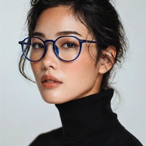 May include: A person wearing round, navy blue eyeglasses with a black turtleneck. The glasses have a thin frame and are positioned on the bridge of the nose. The person has dark hair and freckles.