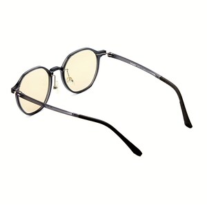 May include: A pair of glasses with translucent dark gray frames and light yellow tinted lenses. The frames feature a rounded shape with a thin bridge and metallic nose pads. The temples are dark gray, transitioning to black tips at the ends.