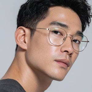 Könnte beinhalten: Eine Person trägt eine goldgerahmte Brille mit quadratischer Form. Die Brille hat einen dünnen Rahmen und klare Gläser. Die Person trägt ein dunkelgraues T-Shirt.