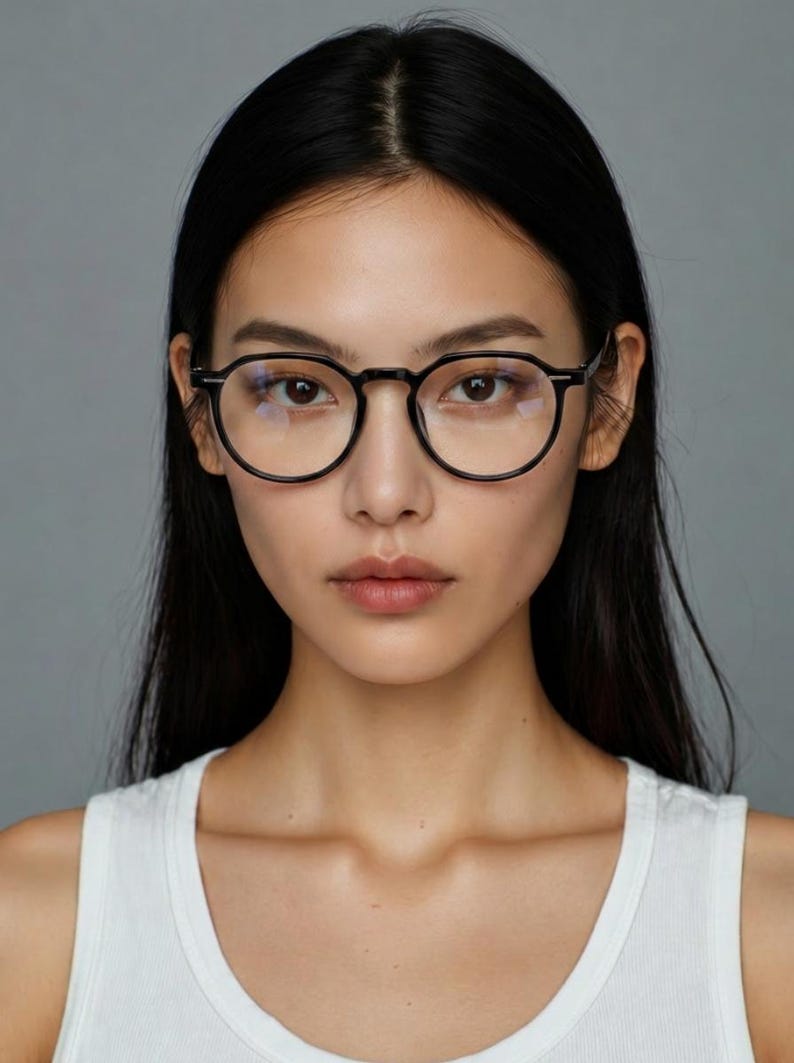 May include: A woman wearing black round-framed eyeglasses. The glasses have a classic design with a thin frame. The woman has long, dark hair and is wearing a white tank top. The background is a solid gray.