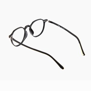 May include: Black eyeglasses with round frames. The glasses have a sleek, modern design with a matte finish. The temples are thin and extend back, and the lenses are clear. The glasses are set against a white background.