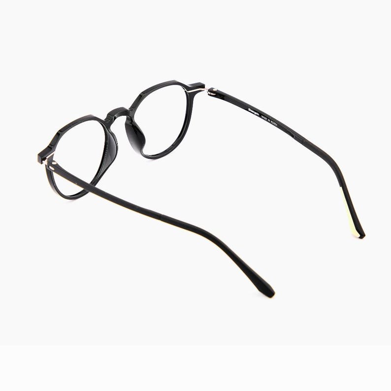 May include: Black eyeglasses with round frames. The glasses have a sleek, modern design with a matte finish. The temples are thin and extend back, and the lenses are clear. The glasses are set against a white background.