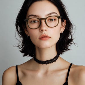 May include: A woman wearing olive green eyeglasses with a square frame and a black lace choker. She is also wearing a black tank top. The background is a solid light gray.
