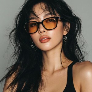 May include: Black-framed sunglasses with orange-tinted lenses. The rectangular frames have a subtle design element on the temples. The model is wearing a black tank top and silver hoop earrings.
